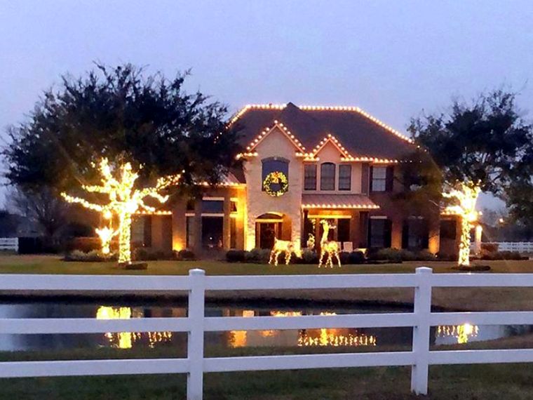 Home with wrapped trees, lit reindeer and roofline lights behind a white fence