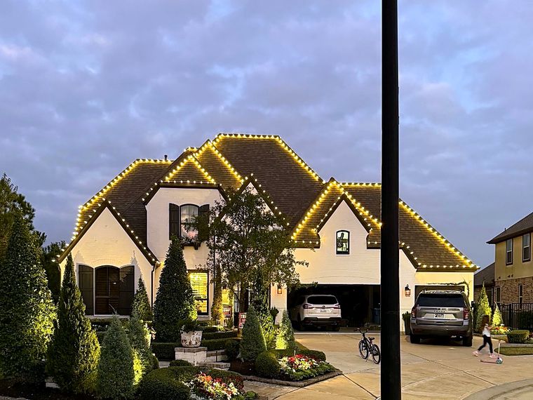Home with warm white roofline lights and lit landscaping at dusk